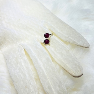 Pode incluir: Um anel de prata com duas pedras redondas de cor vermelho escuro. O anel está sobre uma superfície branca e felpuda.