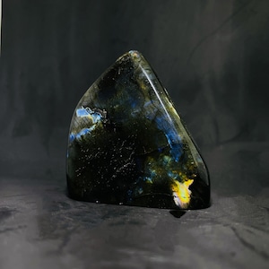 Op de afbeelding: Een donkergrijze, blauwe en gouden labradoritkristal met een driehoekige vorm. De kristal is glad en gepolijst.