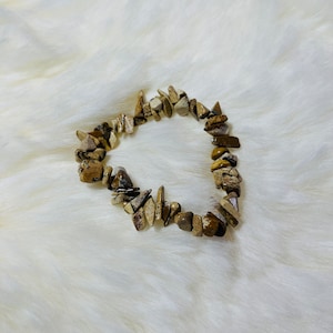 Op de afbeelding: Een bruin en beige stenen armband met verschillende vormen en maten stenen. De armband ligt op een wit, pluche oppervlak.