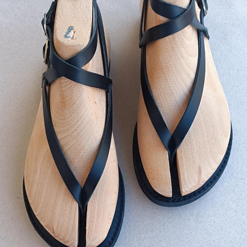 May include: A pair of black leather sandals with a criss-cross strap design. The sandals have a wooden sole and a toe thong.
