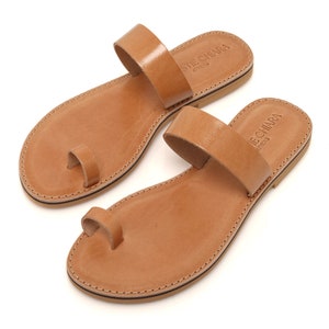 Pode incluir: Um par de sandálias de couro castanho com anel para o dedo do pé e uma única tira na parte superior do pé. As sandálias têm o texto "STIE CHIARA ATHENS" gravado na tira.