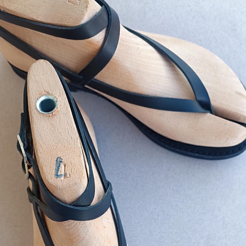 May include: Black leather sandals with a wooden sole and multiple straps. The sandals have a buckle closure and are on a wooden shoe form.