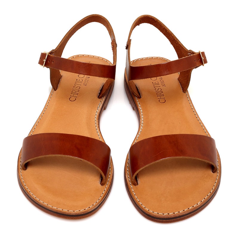 May include: A pair of brown leather sandals with a buckle closure. The sandals have a simple design and are made of high-quality leather. The brand name "Christie C. Athens" is printed on the straps.