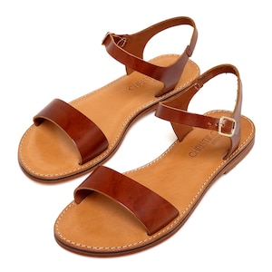 May include: A pair of brown leather sandals with ankle straps and buckles. The sandals have a simple design and are made of high-quality leather. The brand name "Christie  Athens" is printed on the insole.