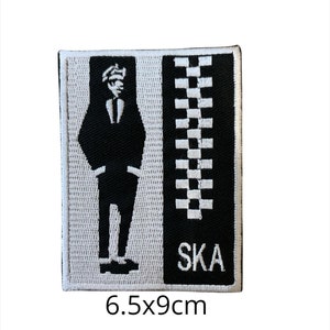 Op de afbeelding: Zwart-witte geborduurde strijkpatch met een silhouet van een persoon in een pak met een geruit patroon en het woord "SKA" in witte tekst. De patch is 6,5 bij 9 centimeter.