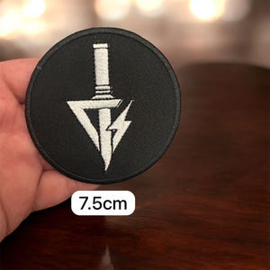 May include: A round, black patch with a white embroidered design. The design features a sword, a downward-pointing triangle, and a lightning bolt. The patch is 7.5cm in diameter.