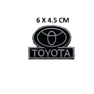 Op de afbeelding: Zwart en wit geborduurde Toyota-patch, afmetingen 6 x 4,5 cm. De patch toont het Toyota-logo in een ovale vorm boven de merknaam. De tekst "TOYOTA" wordt in een rechthoek eronder weergegeven.
