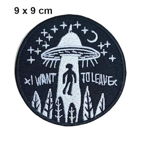 May include: A circular black patch with a white embroidered design. The design includes a UFO, a silhouetted figure, trees, stars, and the text "I WANT TO LEAVE." The patch measures 9 x 9 cm.