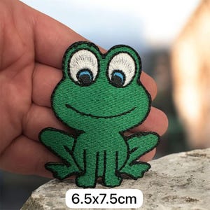Może przedstawiać: Haftowana zielona naszywka z żabą z czarnym konturem i niebieskimi oczami. Naszywka ma wymiary 6,5 x 7,5 cm. Żaba uśmiecha się i ma rozłożone ramiona i nogi. Naszywka znajduje się na jasnej powierzchni.