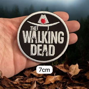May include: A circular black patch with the words "THE WALKING DEAD" in white, and a white hat with a red star. The patch measures 7cm in diameter.
