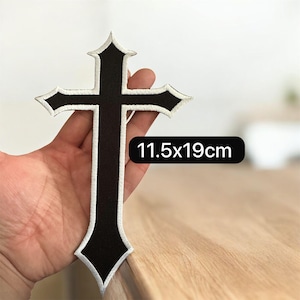 May include: Black embroidered cross patch with white stitching. The patch measures 11.5cm x 19cm. The cross has a decorative design with pointed edges. The patch is held in a hand.