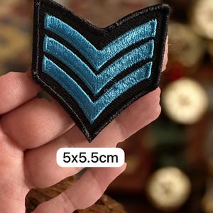 May include: A black embroidered patch with three light blue chevrons. The patch measures 5 x 5.5 cm. The patch is held in a hand.