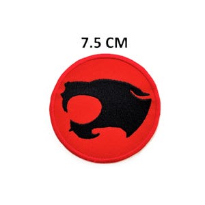 Może przedstawiać: Okrągła, czerwona naszywka z czarną grafiką stylizowanej głowy kota. Naszywka ma średnicę 7,5 cm. Naszywka ma obszytą krawędź i prawdopodobnie jest to emblemat do naprasowywania lub przyszywania.