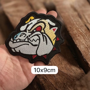 May include: Embroidered patch featuring a bulldog's head with a fierce expression. The design incorporates white, yellow, red, and black colors. The patch measures approximately 3.9 x 3.5 inches, as indicated by the text on the image.