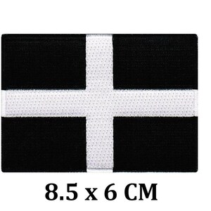 Könnte beinhalten: Schwarz-weißes Flaggen-Patch mit einem weißen Kreuz auf schwarzem Hintergrund. Das Patch misst 8,5 x 6 cm.