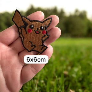 Puede incluir: Un parche bordado marrón con un personaje de dibujos animados con contornos negros, mejillas rojas y una boca sonriente. El parche mide 6x6cm. El parche se sostiene en una mano sobre un fondo verde borroso.