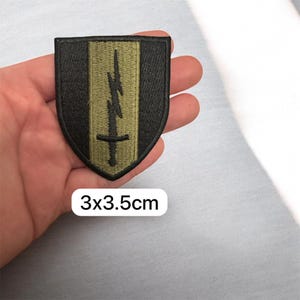 Könnte beinhalten: Ein schwarz-olivgrüner, gestickter Aufnäher in Schildform. Das Design zeigt einen Blitz über einem Schwert. Der Aufnäher misst 3 x 3,5 cm.