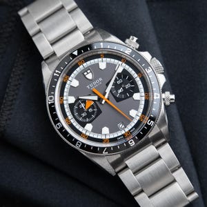 May include: A stainless steel Tudor Geneve chronograph watch with a grey dial, black bezel, and orange accents. The watch features a metal link bracelet, two black sub-dials, a date window at the six o'clock position, and luminous hour markers.