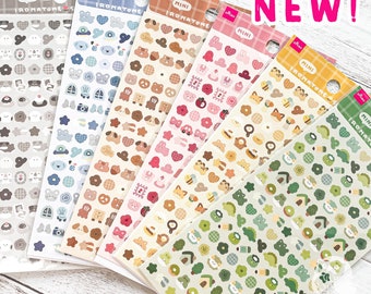 May include: Six sheets of colorful mini stickers with various cute animal and object designs. The stickers are arranged in a fan shape on a white wooden surface. The text "NEW!" is written in pink letters on the top right corner of the image.
