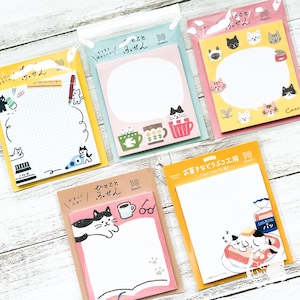 Może przedstawiać: Kolekcja karteczek samoprzylepnych z motywem kota, każda zawierająca 30 kartek. Karteczki mają różne wzory, w tym koty, filiżanki do kawy i ilustracje jedzenia. Karteczki są zapakowane w przezroczysty plastik.