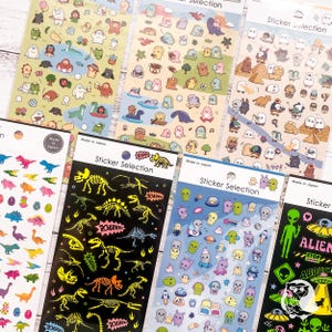 Può includere: Una collezione di fogli di adesivi con vari disegni. I fogli includono dinosauri colorati, personaggi dei cartoni animati e adesivi a tema alieno. Alcuni fogli hanno il testo "Sticker Selection" e "Made in Japan".