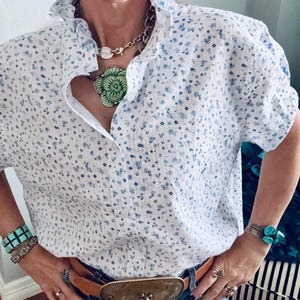 May include: White blouse with a blue floral pattern and ruffled collar. The blouse is paired with a brown leather belt with a large, ornate buckle. Accessories include turquoise bracelets, earrings, and a necklace with a green pendant. The model is wearing blue jeans.