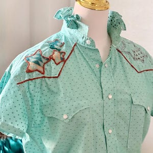 May include: A light turquoise short-sleeve shirt with a dotted pattern and star-shaped appliqu&eacute;s. The shirt features a ruffled collar, button-down front, and two chest pockets. The star appliqu&eacute;s have a red outline and are filled with a geometric pattern.
