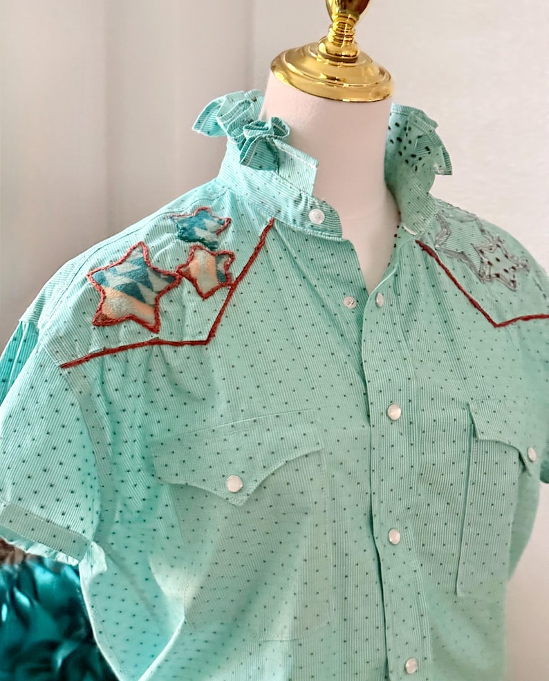 May include: A light turquoise short-sleeve shirt with a dotted pattern and star-shaped appliqu&eacute;s. The shirt features a ruffled collar, button-down front, and two chest pockets. The star appliqu&eacute;s have a red outline and are filled with a geometric pattern.