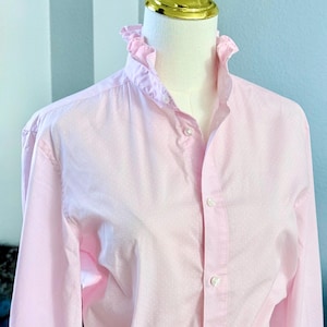 Könnte beinhalten: Ein hellrosa Hemd mit Rüschenkragen und kleinen weißen Tupfen. Das langärmlige Hemd wird auf einer Schaufensterpuppe präsentiert. Das Hemd hat ein klassisches Design.