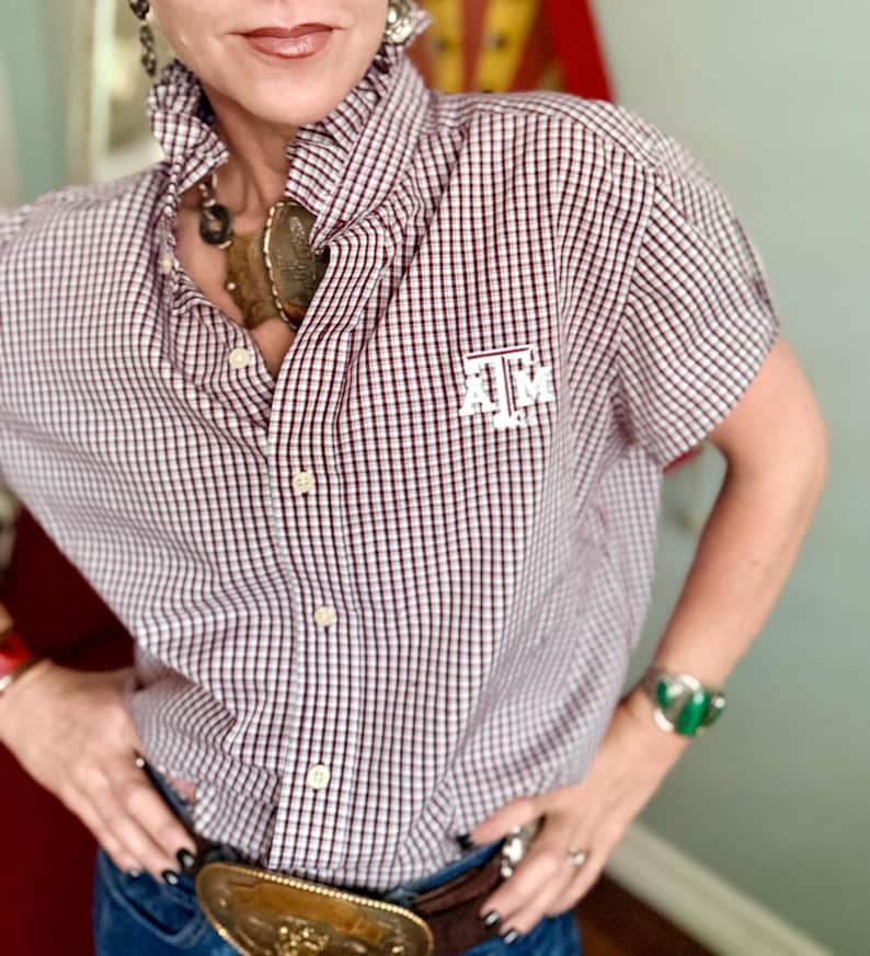 Peut inclure: Une chemise &agrave; manches courtes &agrave; carreaux rouges et blancs. La chemise porte les lettres "A M" brod&eacute;es sur la poitrine. Elle est port&eacute;e avec un jean bleu et une ceinture marron &agrave; boucle dor&eacute;e. Un style classique.