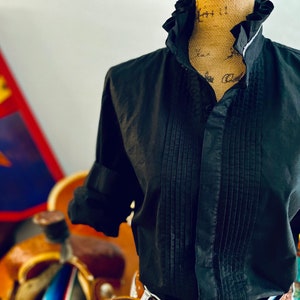 Peut inclure: Chemise noire boutonn&eacute;e avec col &agrave; volants et devant pliss&eacute;. La chemise a des manches longues avec des poignets retrouss&eacute;s. La chemise est pr&eacute;sent&eacute;e sur un mannequin. L'arri&egrave;re-plan comprend une pi&egrave;ce d&eacute;corative color&eacute;e.