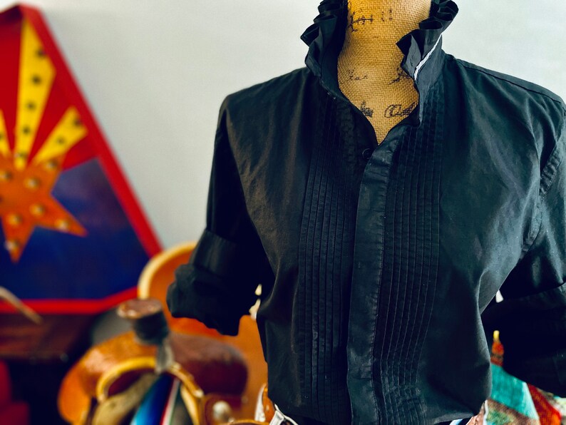 Peut inclure: Chemise noire boutonn&eacute;e avec col &agrave; volants et devant pliss&eacute;. La chemise a des manches longues avec des poignets retrouss&eacute;s. La chemise est pr&eacute;sent&eacute;e sur un mannequin. L'arri&egrave;re-plan comprend une pi&egrave;ce d&eacute;corative color&eacute;e.