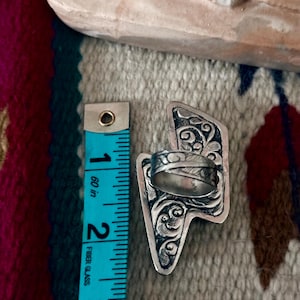May include: A silver-toned ring with an ornate, engraved design. The ring's decorative element is approximately 2.5 inches long, as measured by a blue measuring tape. The ring is set against a patterned textile background.