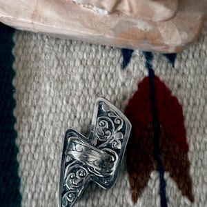 May include: A silver-colored ring in the shape of a lightning bolt, featuring intricate floral and swirl patterns. The ring is set against a textured, patterned fabric background, with a hint of a light-colored sculpture in the upper portion of the image.