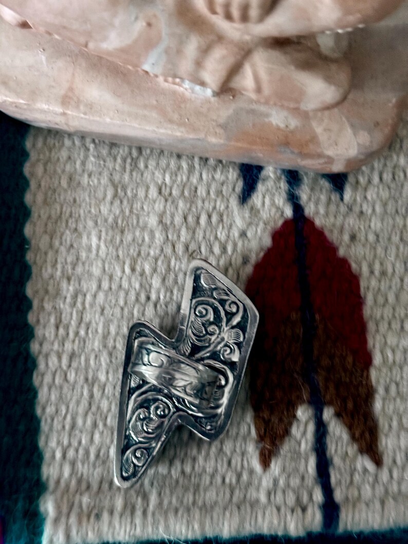 May include: A silver-colored ring in the shape of a lightning bolt, featuring intricate floral and swirl patterns. The ring is set against a textured, patterned fabric background, with a hint of a light-colored sculpture in the upper portion of the image.