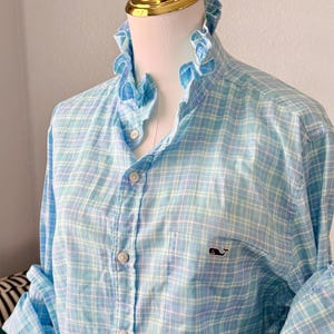 May include: A long-sleeved button-down shirt in a light blue, white, and pale green plaid pattern. The shirt features a ruffled collar, a front button placket, and a single chest pocket with a small embroidered whale logo in dark navy blue.