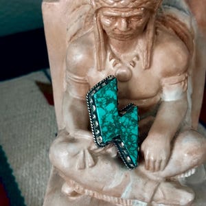 May include: A turquoise and silver-toned lightning bolt pendant. The turquoise stone has a marbled appearance and is set in a silver-toned metal frame with a beaded edge. The pendant is displayed on a statue.