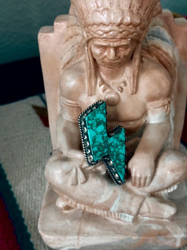 May include: A turquoise and silver-toned lightning bolt pendant. The turquoise stone has a marbled appearance and is set in a silver-toned metal frame with a beaded edge. The pendant is displayed on a statue of a Native American.