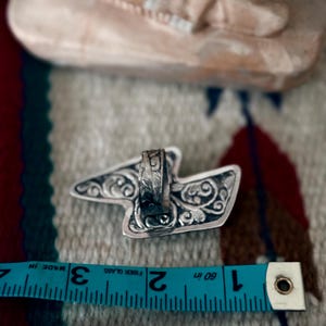 May include: A silver ring with an ornate, stylized design. The ring's decorative elements include swirling patterns. A blue measuring tape shows the ring is approximately 2 inches long.