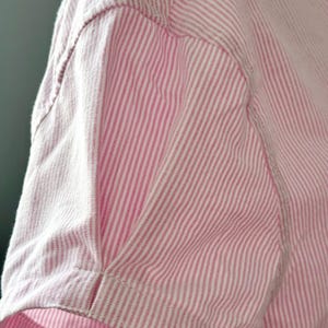 Peut inclure: Gros plan sur une chemise &agrave; manches courtes ray&eacute;e rose et blanche. Le tissu pr&eacute;sente un motif &agrave; rayures verticales. La manche est cousue avec des coutures visibles.