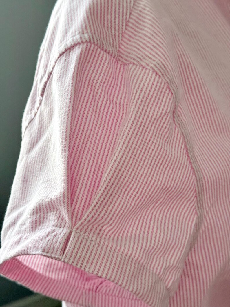Peut inclure: Gros plan sur une chemise &agrave; manches courtes ray&eacute;e rose et blanche. Le tissu pr&eacute;sente un motif &agrave; rayures verticales. La manche est cousue avec des coutures visibles.
