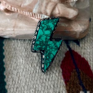 May include: A turquoise-colored lightning bolt-shaped pendant with a silver-colored rope-like border. The turquoise has dark green veining. The pendant is set against a textured, off-white background.