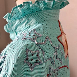 May include: A turquoise and white striped shirt with a ruffled collar and star-shaped patches. The shirt has a dotted pattern and red stitching accents. The stars are outlined with a denim-like material.