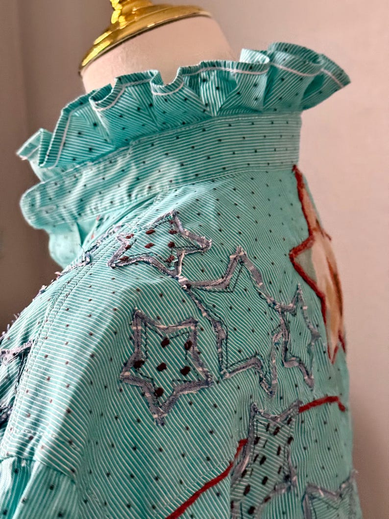 May include: A turquoise and white striped shirt with a ruffled collar and star-shaped patches. The shirt has a dotted pattern and red stitching accents. The stars are outlined with a denim-like material.