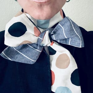 May include: A patterned scarf with a bow tied at the neck. The scarf features a mix of white, navy, and light blue stripes and large polka dots in navy, brown, and red. The scarf is worn over a dark blue jacket.