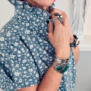 Peut inclure: Un chemisier bleu &agrave; imprim&eacute; floral avec un col &agrave; volants et des manches courtes. La personne porte plusieurs bagues, un bracelet manchette en argent avec des pierres vertes et des boucles d'oreilles roses.
