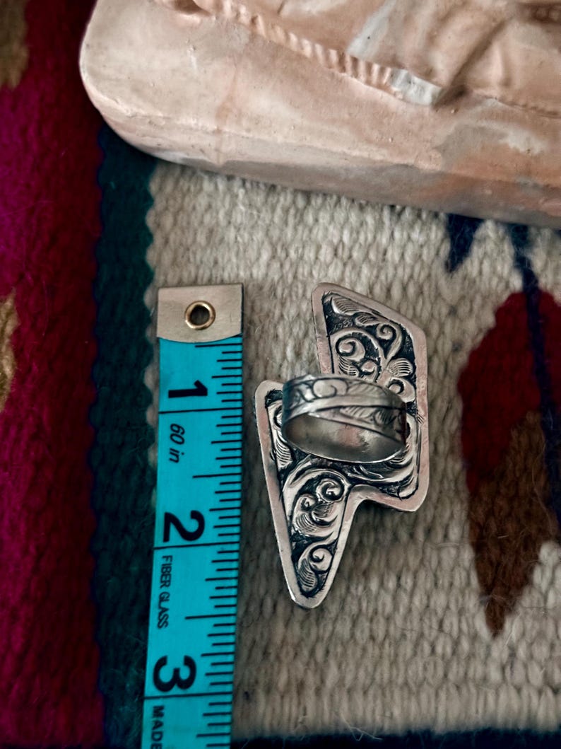 May include: A silver ring with an ornate, engraved design. The ring is approximately 2.5 inches long, as measured by a blue measuring tape. The ring's design features swirling patterns and is set against a patterned fabric.