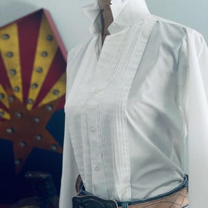 Peut inclure: Une chemise blanche &agrave; col volant&eacute; et devant pliss&eacute;. La chemise est associ&eacute;e &agrave; une ceinture marron clair et une boucle d&eacute;corative argent&eacute;e. La chemise est rentr&eacute;e dans un jean bleu.