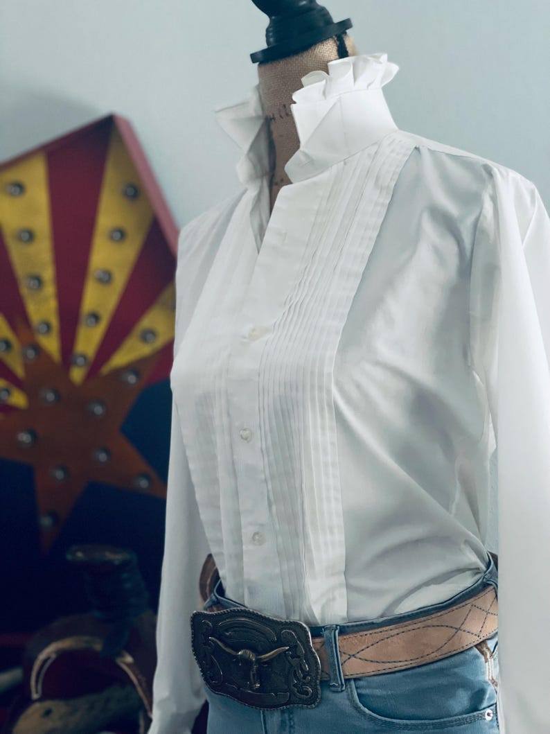 Peut inclure: Une chemise blanche &agrave; col volant&eacute; et devant pliss&eacute;. La chemise est associ&eacute;e &agrave; une ceinture marron clair et une boucle d&eacute;corative argent&eacute;e. La chemise est rentr&eacute;e dans un jean bleu.