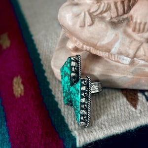 May include: A silver ring with a turquoise stone in a lightning bolt shape. The turquoise is a vibrant green-blue color. The ring has a detailed, textured design around the stone and on the band.